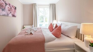 Una cama bien hecha con una colcha a cuadros, almohadas blancas y almohadas rojas estampadas. Hay una mesita de noche con una lámpara y un florero con una flor. Se ve una ventana con cortinas al fondo.