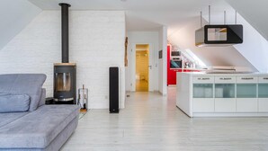 Ein modernes Wohnzimmer mit einem grauen Sofa, einem schwarzen Kaminofen und einer weißen Küche mit einem roten Gerät.