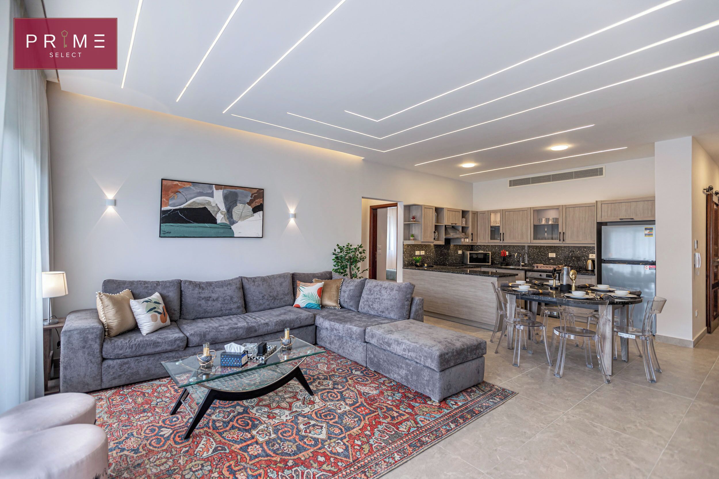 A modern living room with a grey sectional sofa, a glass coffee table, and a patterned rug. A dining area with a glass table and chairs is visible in the background.