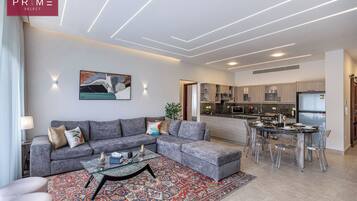 A modern living room with a grey sectional sofa, a glass coffee table, and a patterned rug. A dining area with a glass table and chairs is visible in the background.
