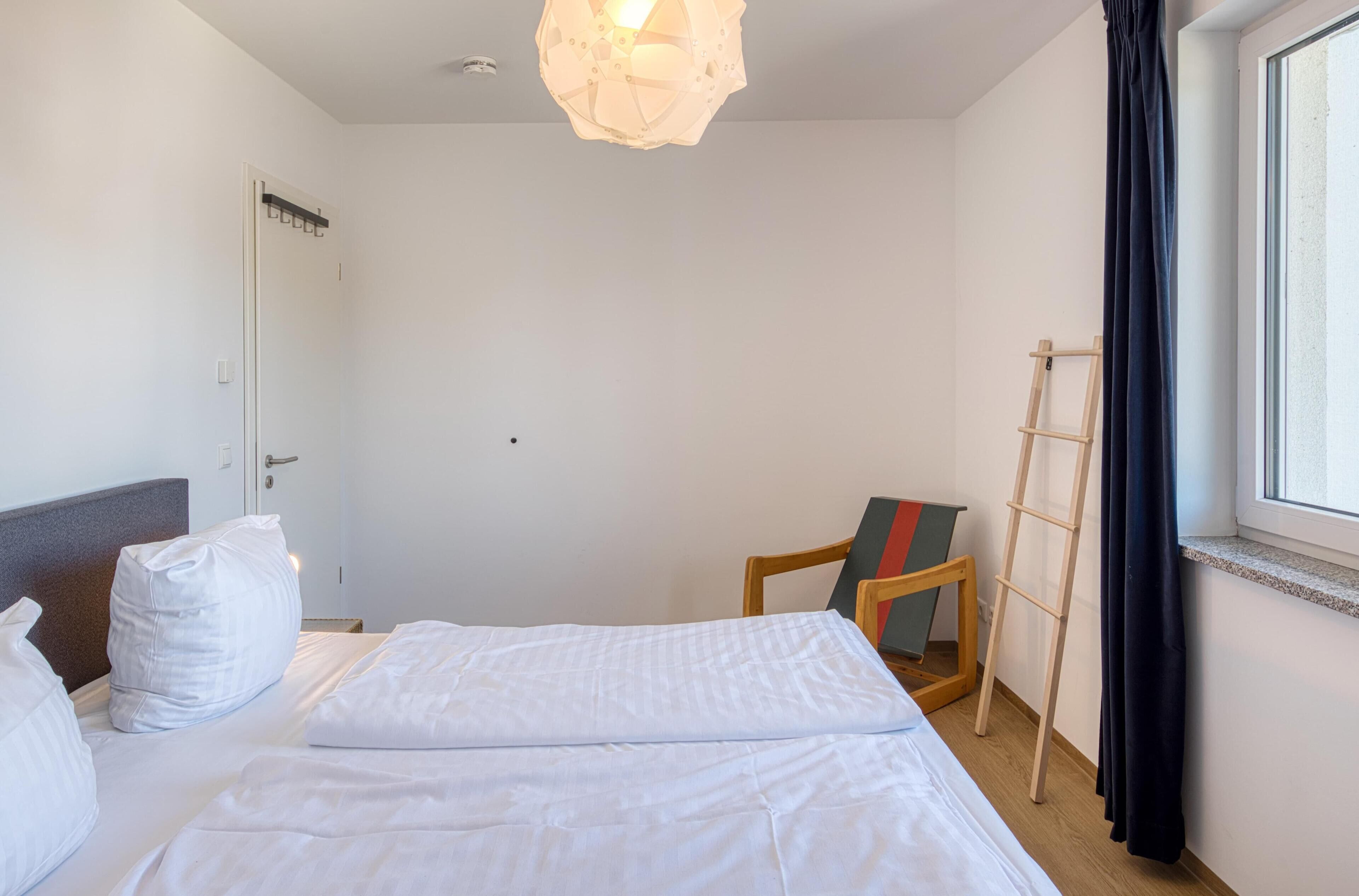 Habitación de hotel con cama, ropa de cama blanca, lámpara colgante, ventana con cortinas y escalera de madera.