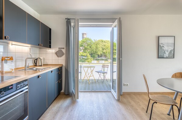 Una cocina moderna con gabinetes grises, un lavamanos de acero inoxidable y un horno empotrado. Una puerta corrediza de vidrio da a una zona de estar exterior con una mesa y sillas.