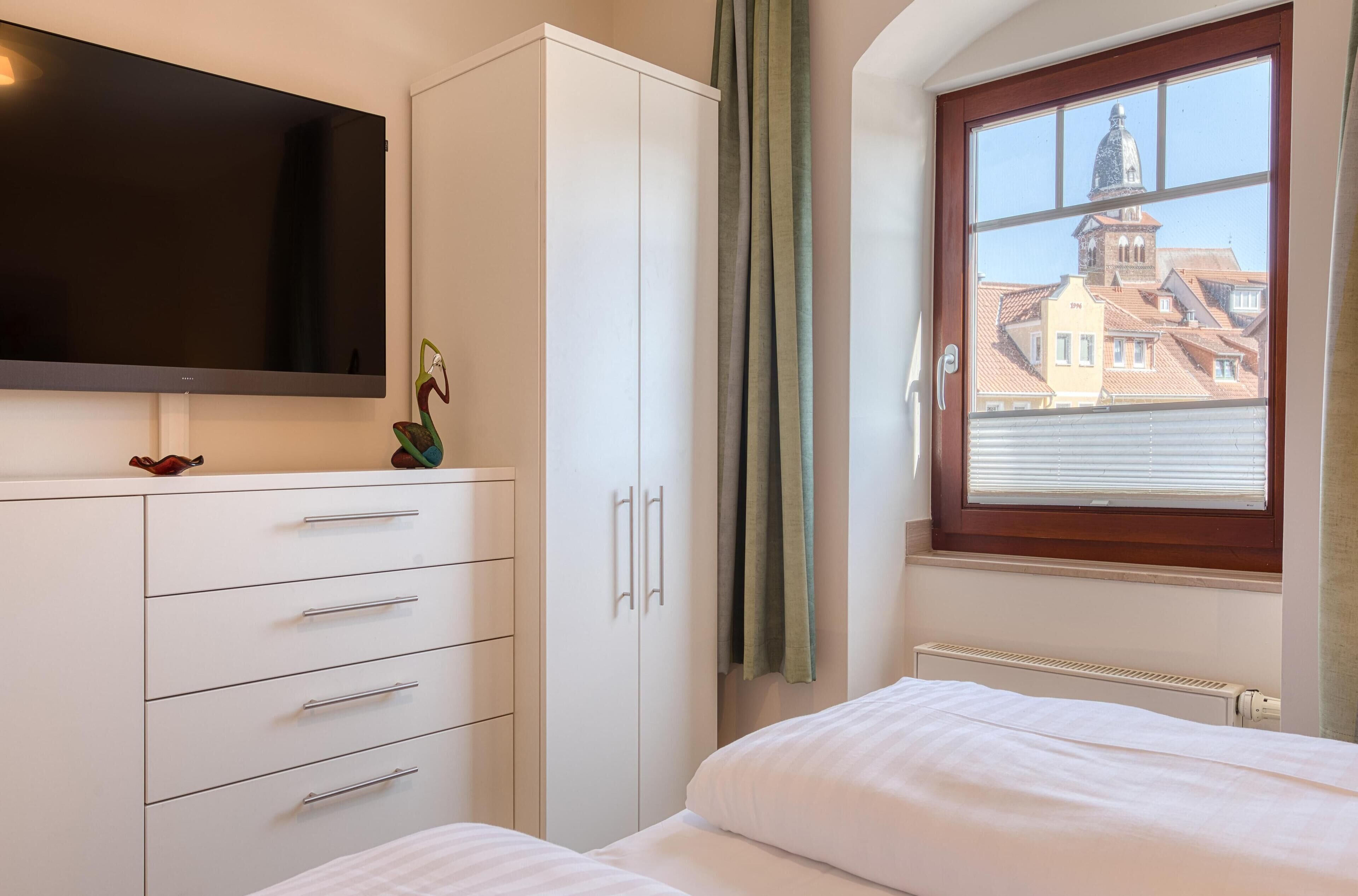 Un dormitorio con cama, armario, televisor de pantalla plana, ventana con vista a edificios y radiador.
