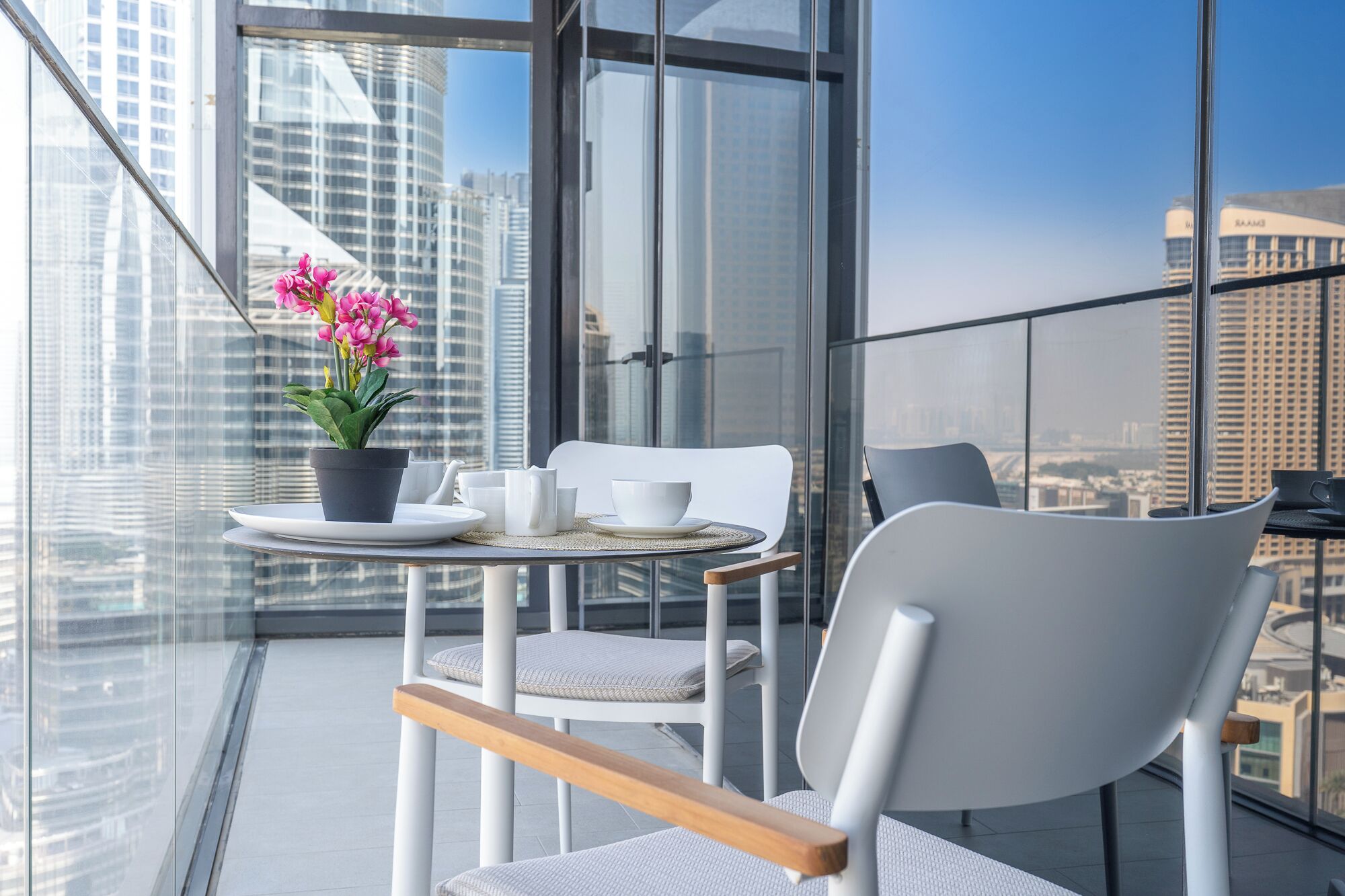 Un balcón moderno con barandilla de vidrio, una mesa blanca con planta en maceta y vistas de la ciudad.
