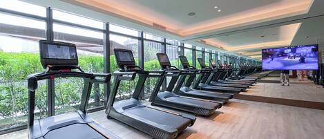 A row of treadmills with screens, large windows, and a TV screen in a gym.