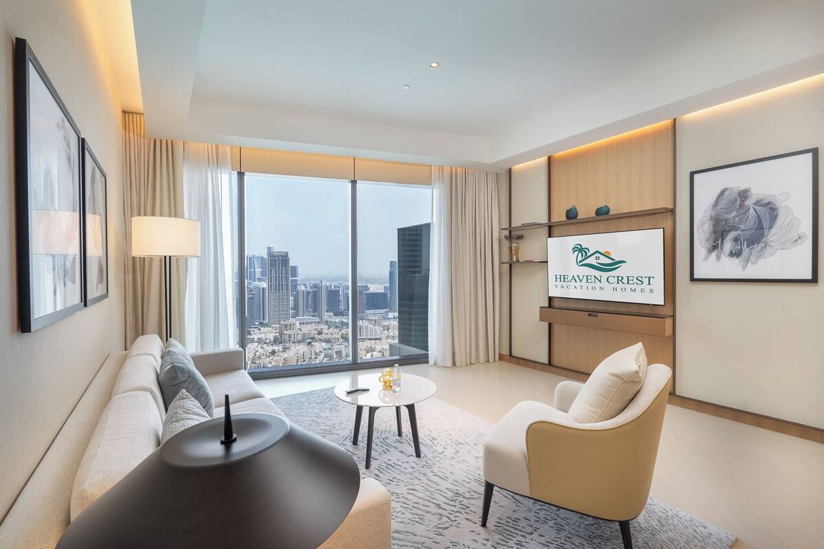 Une chambre d’hôtel moderne dotée d’une grande fenêtre donnant sur la ville, d’un canapé, d’un fauteuil, d’une petite table et d’un écran fixé au mur qui affiche le logo de l’hôtel.