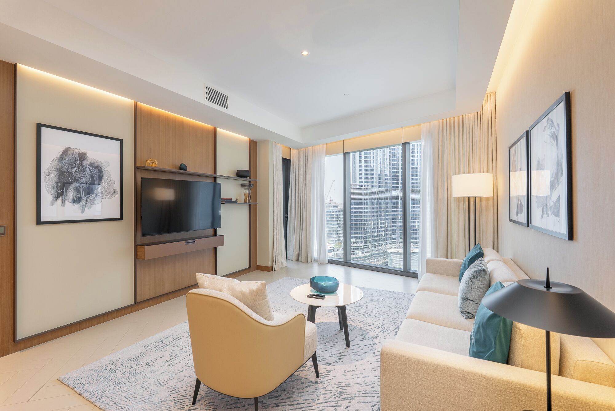 A modern hotel room with a large TV, comfortable seating, a dining area, and a view of the cityscape.