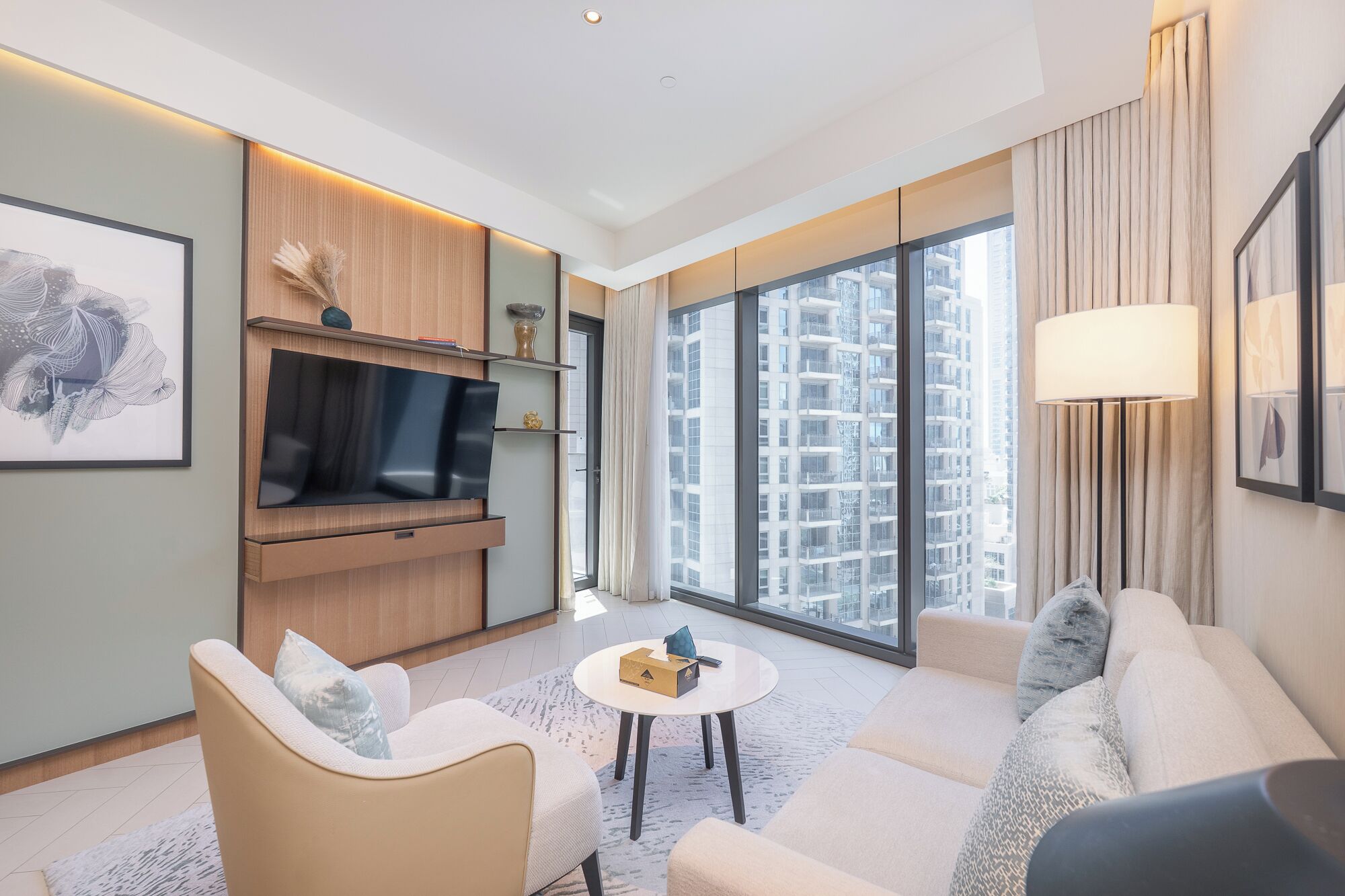 A modern living room with a flat-screen TV, comfortable seating, a small table, and large windows offering a city view.