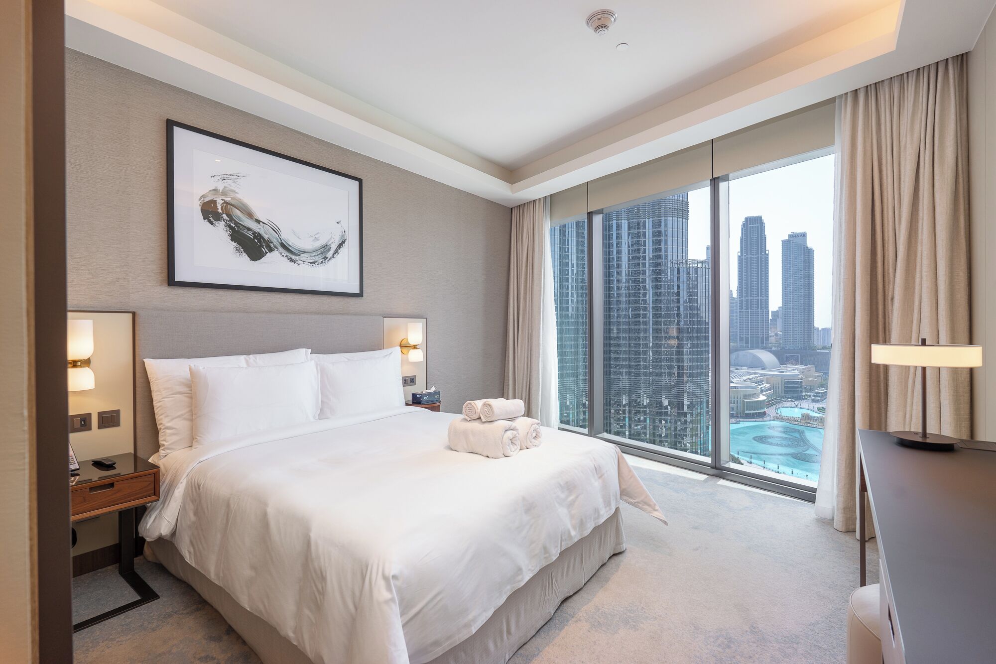 A hotel room with a large bed, a city view, and a framed artwork on the wall.