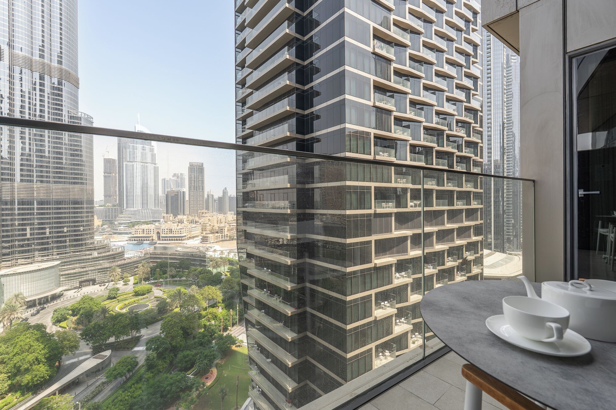 Un moderno edificio de gran altura con fachada de vidrio, un balcón con mesa y sillas, y vistas a un paisaje urbano con otros edificios y zonas verdes.