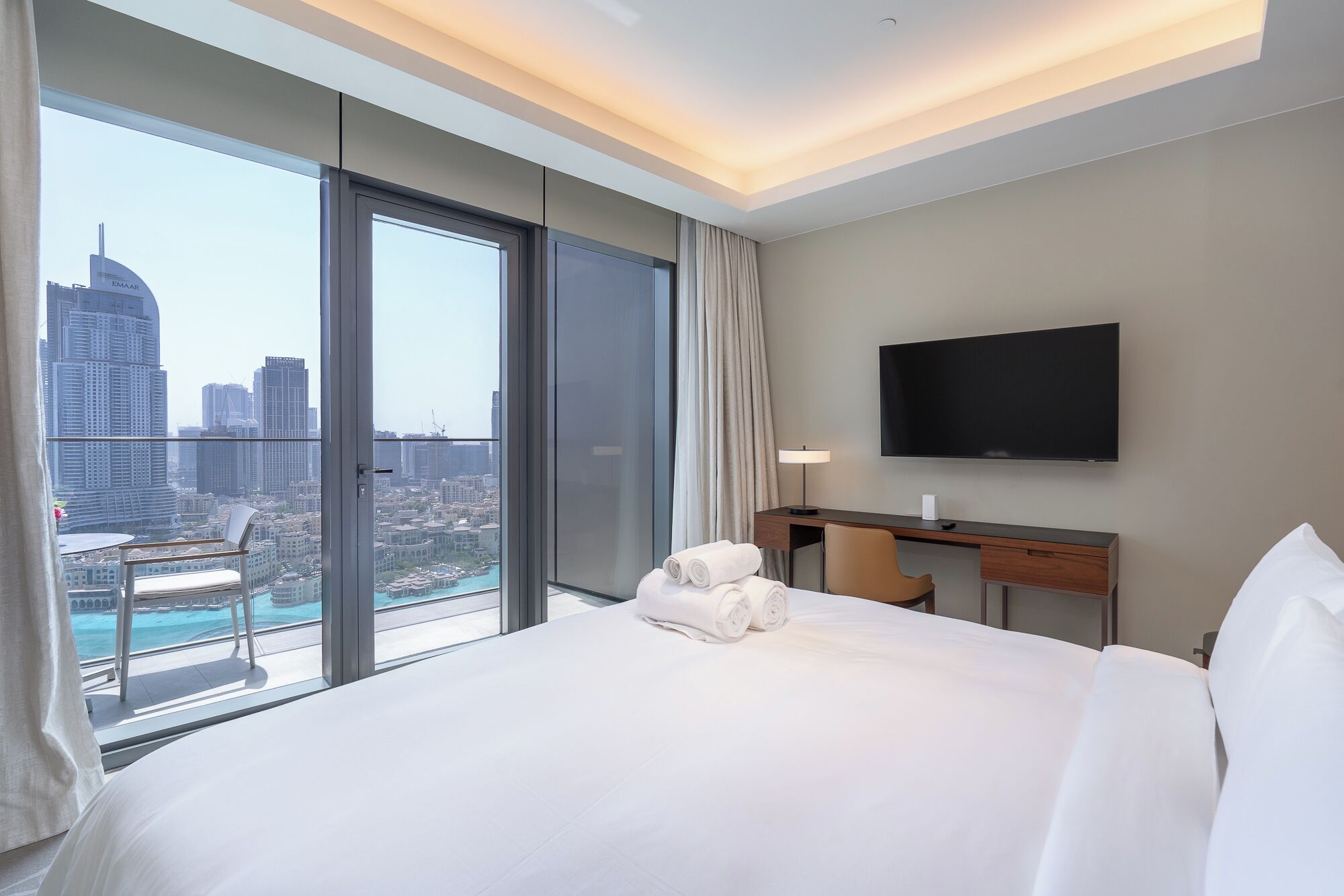 A hotel room with a large bed, a desk, a chair, a TV, and a view of a cityscape through a large window.