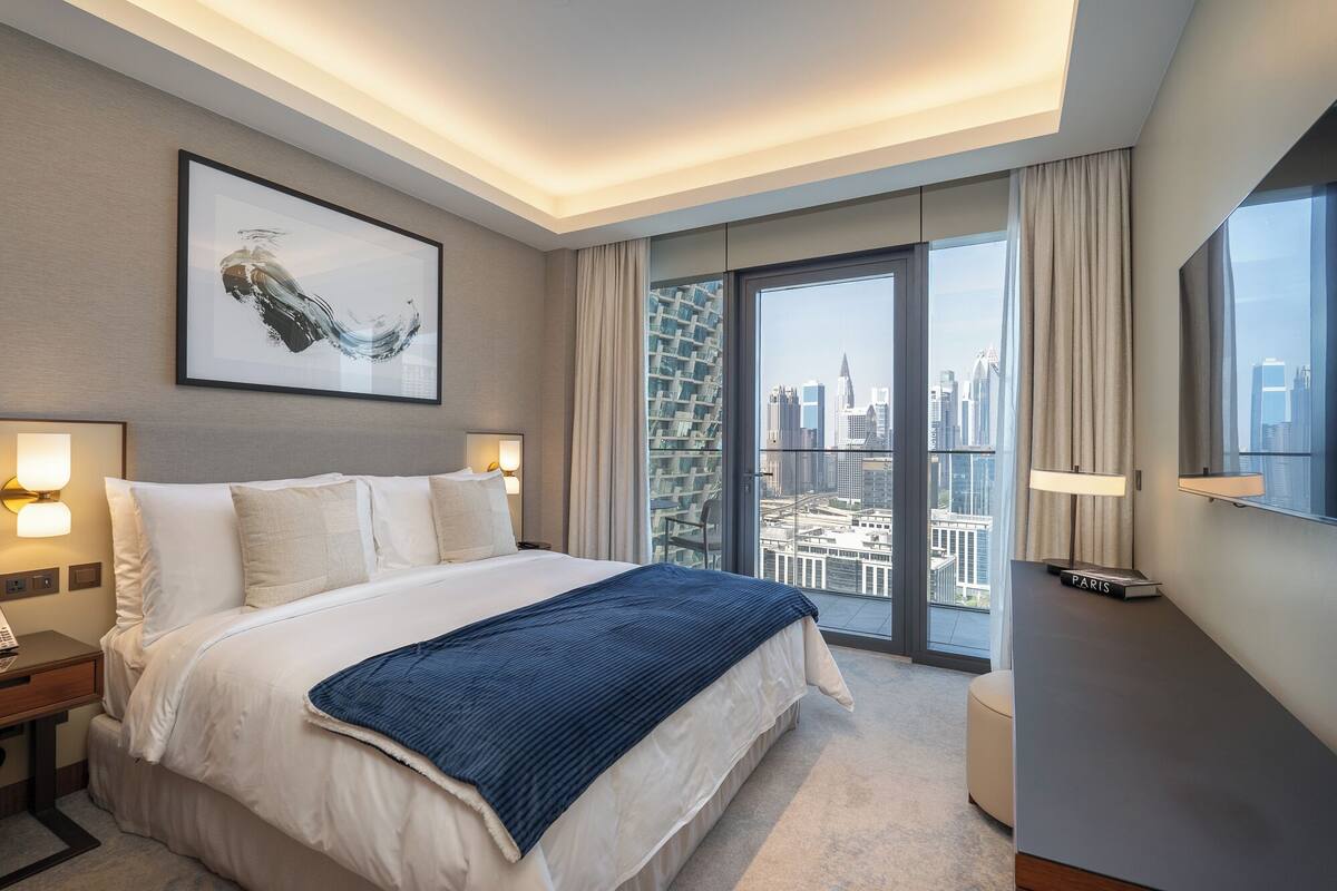 Une chambre d’hôtel avec un grand lit, une vue sur la ville à travers une porte vitrée, une œuvre d’art encadrée au mur et une table de chevet avec une lampe.