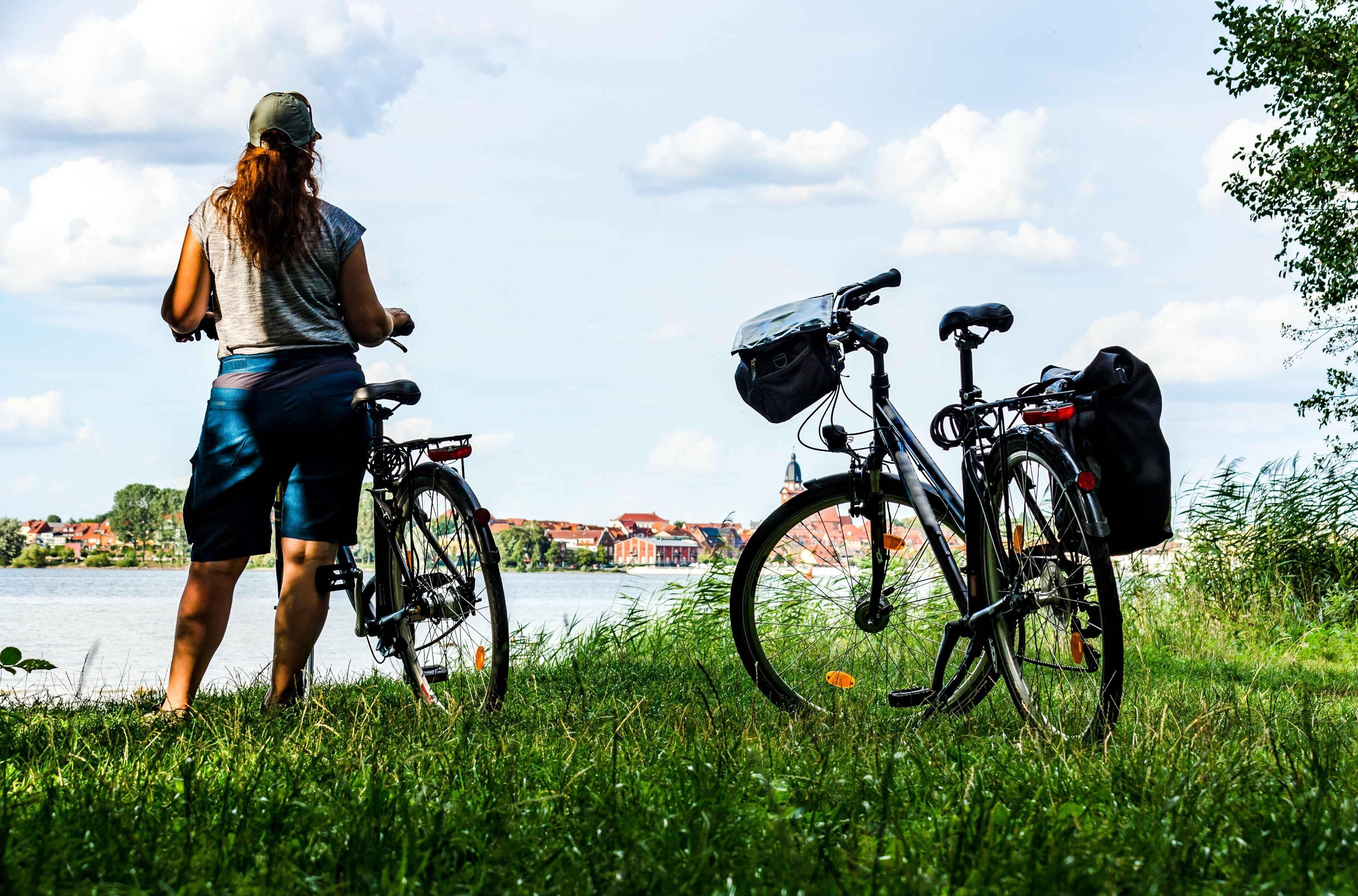 A person standing next to a bicycle with a bag attached, overlooking a body of water with buildings in the background.