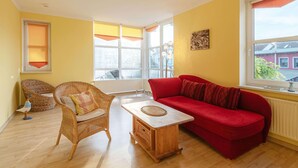 Ein helles Wohnzimmer mit gelben Wänden, einem roten Sofa, Korbstühlen und einem Holztisch.