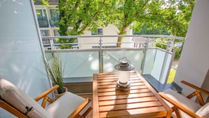 Een balkon met een houten tafel en stoelen, een bloempot en uitzicht op een gebouw met balkons.