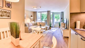 Een moderne woonkamer met een bank, salontafel en eettafel. Er is een groot raam met gordijnen, een plant en uitzicht op buiten.