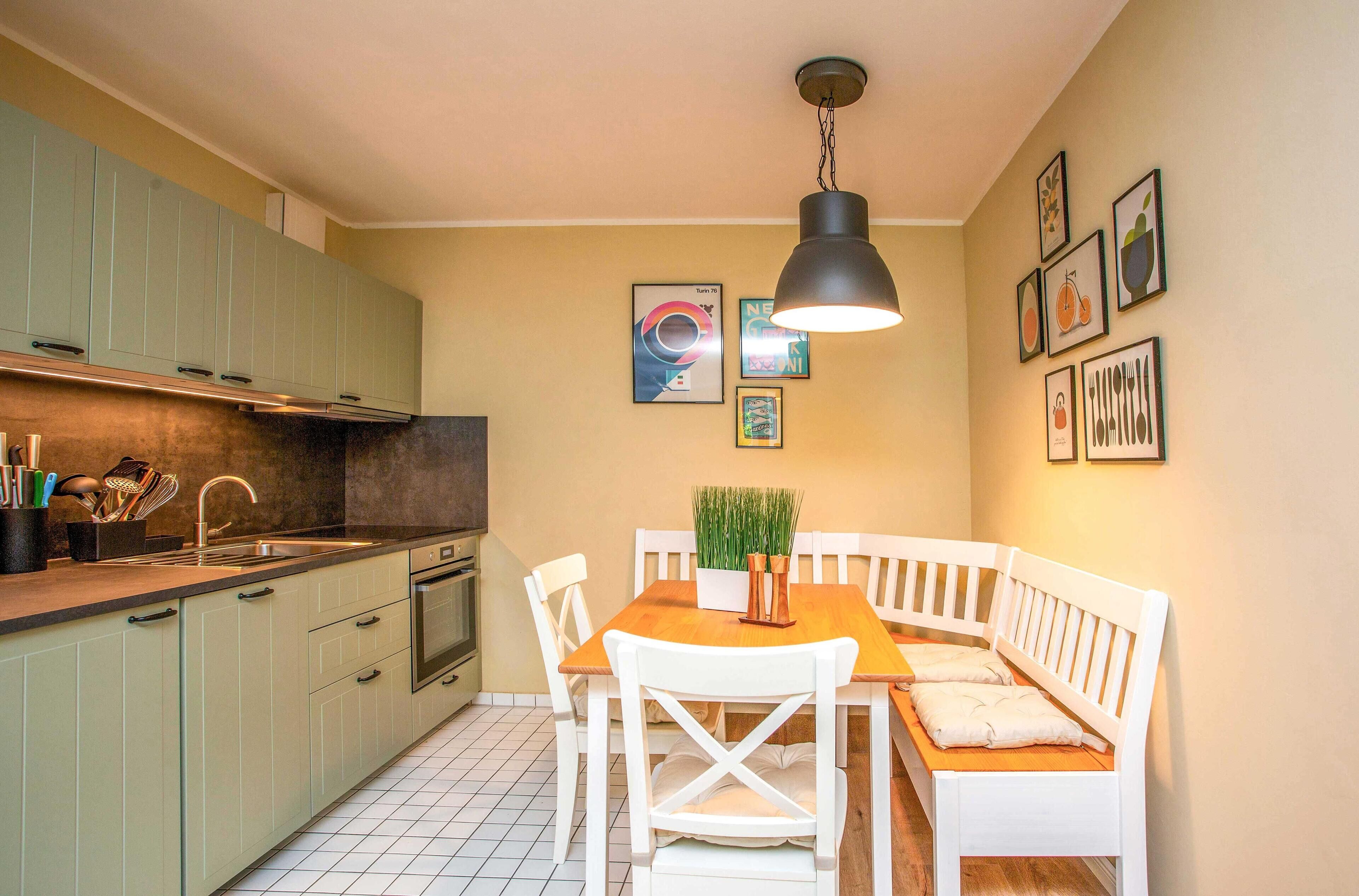A compact kitchen with a dining area, featuring a wooden table and white chairs, a hanging light fixture, and various framed artworks on the walls.