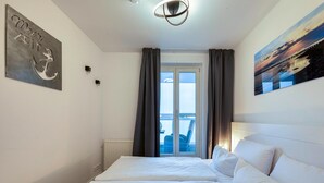 A hotel room with a bed, a view of the sea through a window, and a painting on the wall. - 5-Star "Ostseeperle" Vacation Apartment in Zingst, Next to the Pier (Zingst)