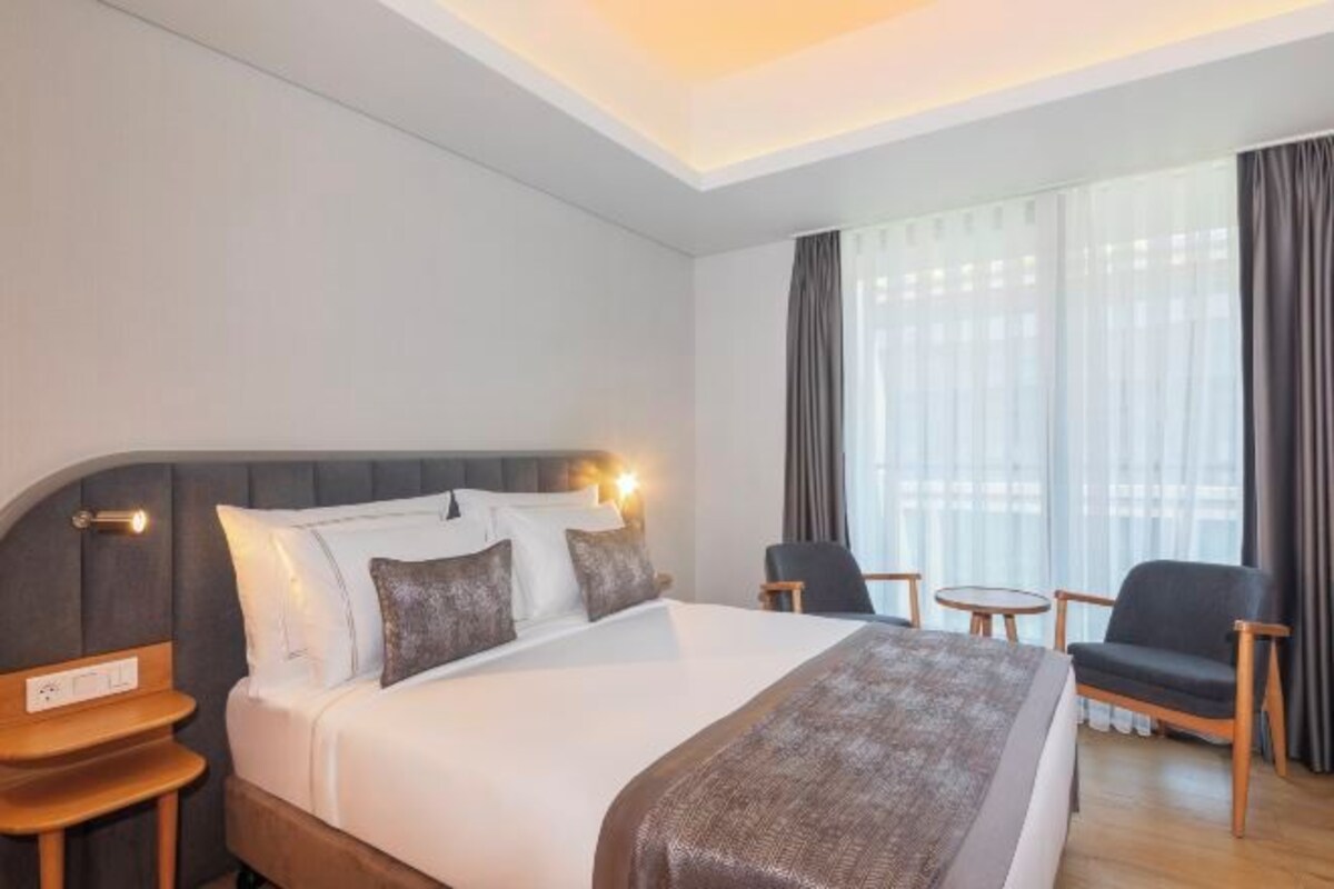A hotel room with a large bed, a bedside table, a chair, a small table, and a window with curtains.