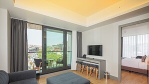 A modern living room with a sofa, ottoman, and a desk with a TV. Large windows offer a city view.