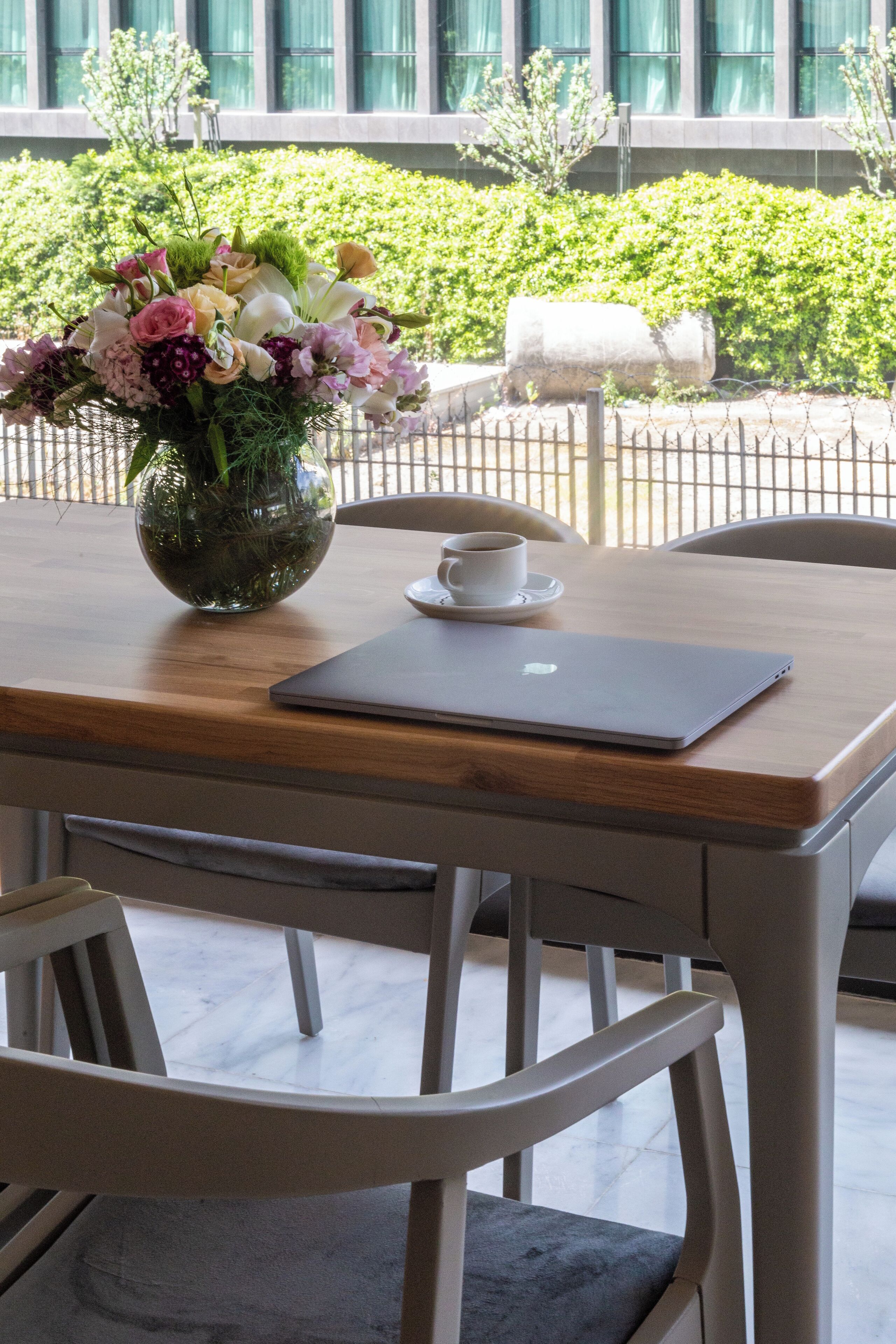 Una mesa de madera con un ordenador portátil, una taza blanca y un jarrón de flores.