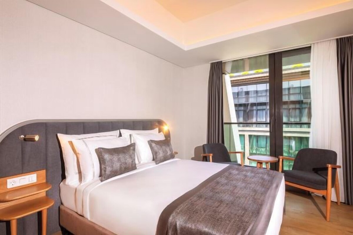 A hotel room with a large bed, a bedside table, a chair, a small table, and a window with curtains.