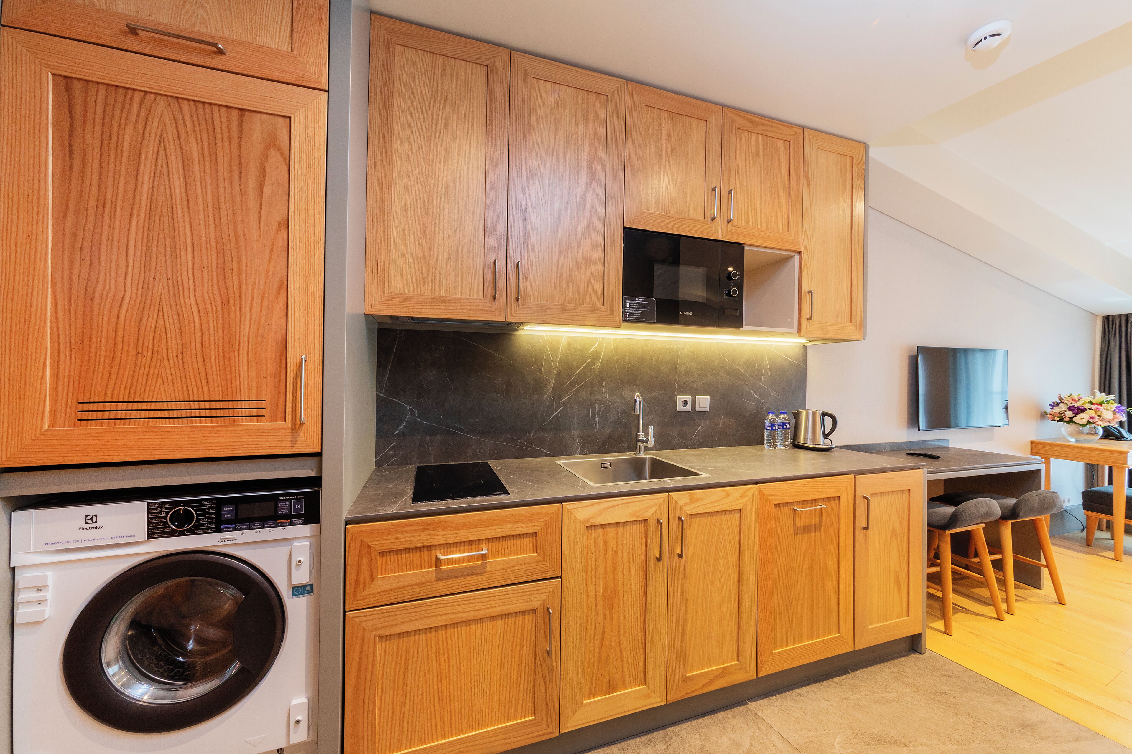 A modern kitchen with wooden cabinets, a built-in microwave, a washing machine, and a dining area with stools.