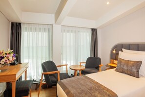 A hotel room with a bed, two chairs, a small table, a desk, and a lamp.