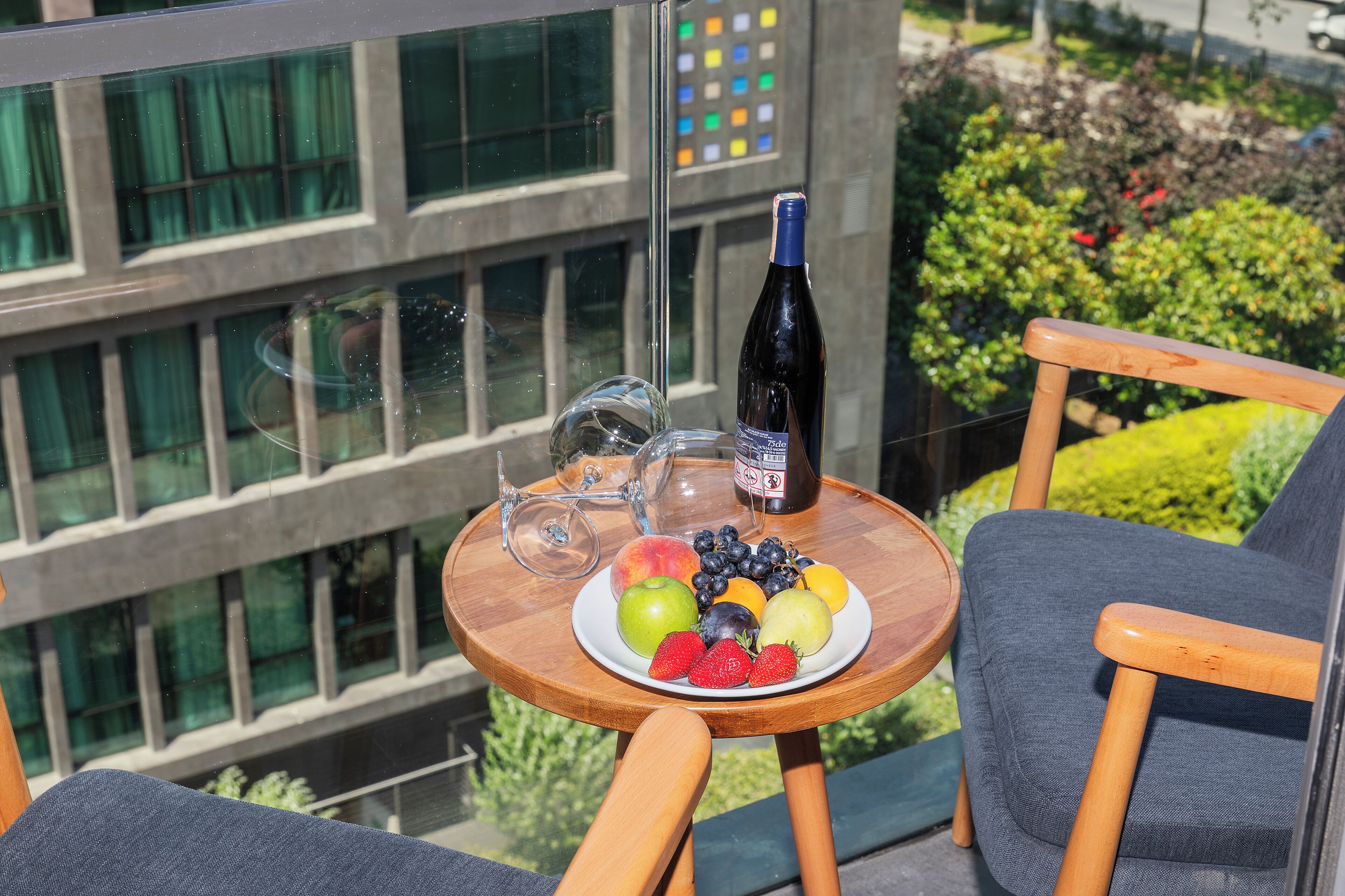 A round wooden table with a plate of fruit, a bottle of wine, and two glasses on a balcony overlooking a building with large windows and greenery.