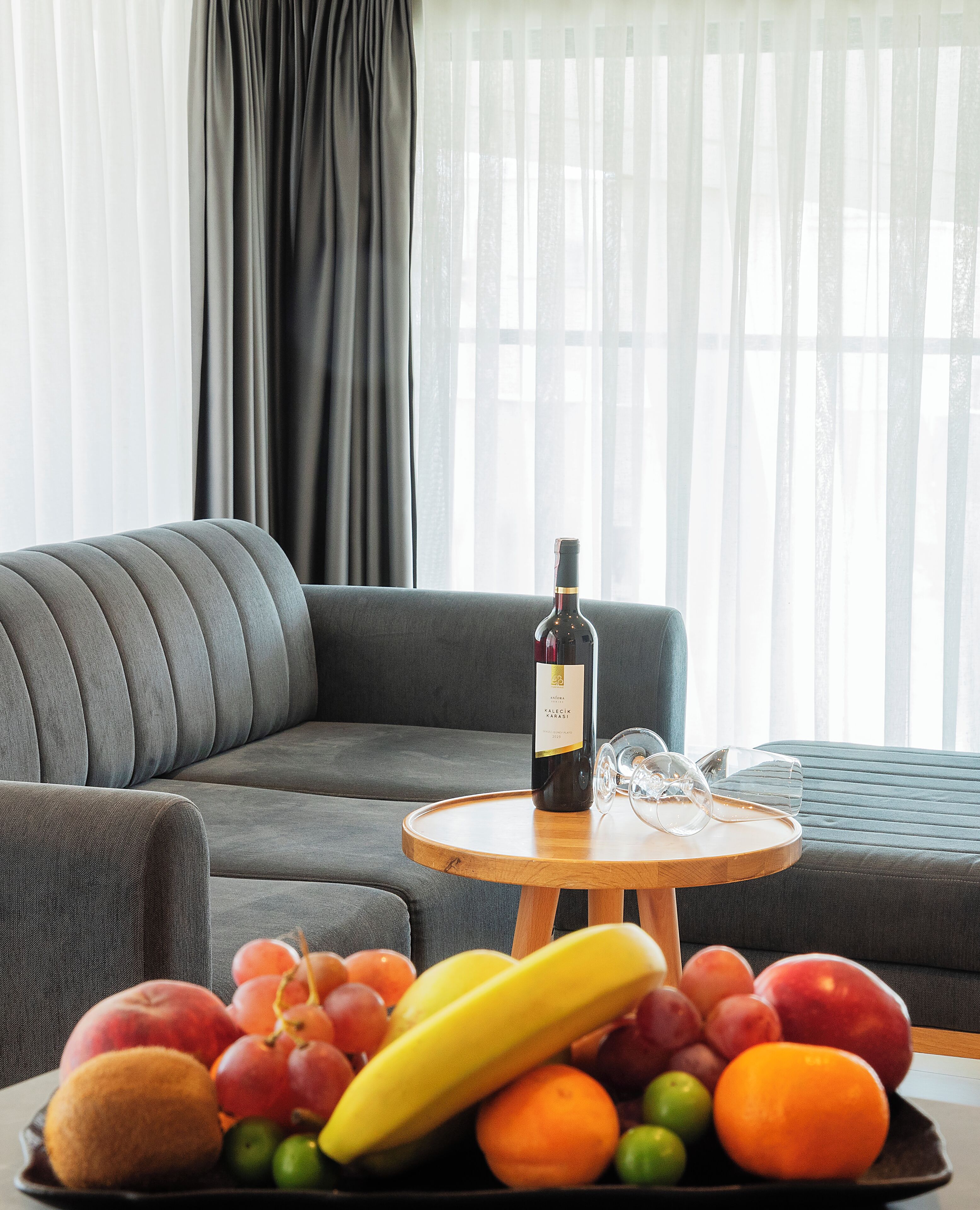 A room with a grey sofa, a round wooden table with a bottle of wine and glasses, and a fruit bowl with various fruits.
