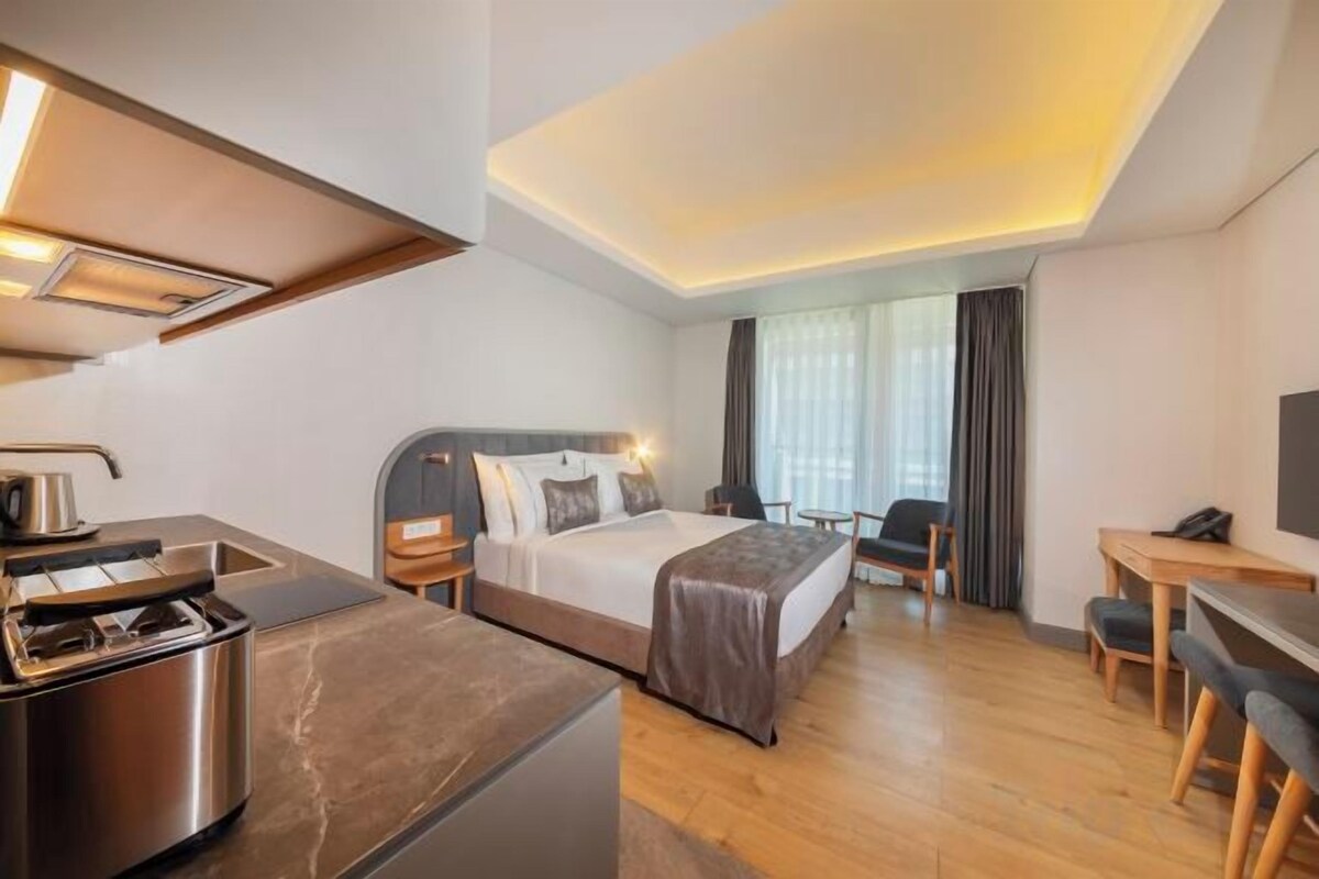A hotel room with a bed, a desk, a chair, a kitchenette, and a large window.