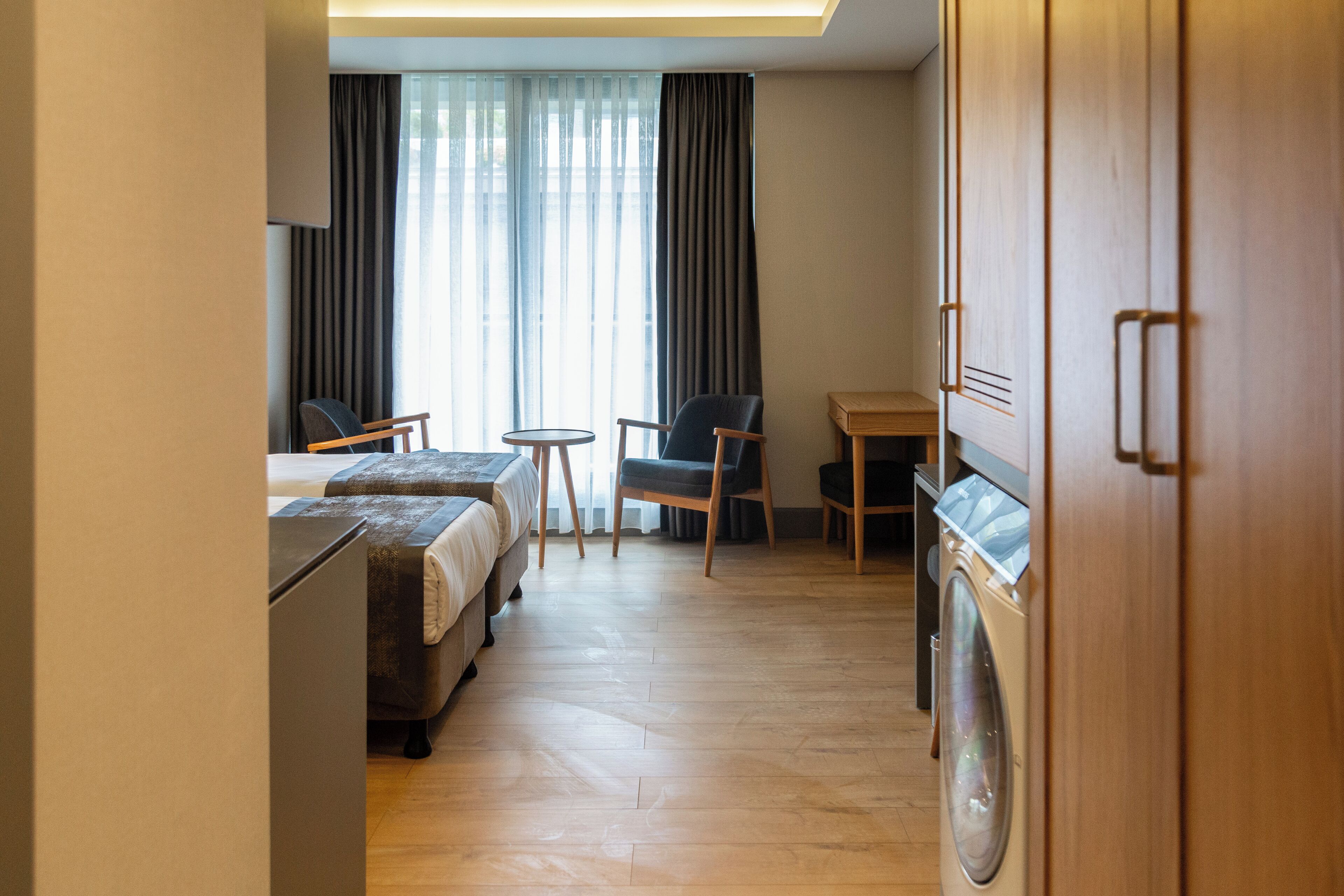 Hotelkamer met twee bedden, een bureau, een stoel, een wasmachine en een groot raam met gordijnen.