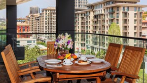 A round wooden table with chairs, a vase of flowers, plates, cups, and food items on a balcony with a view of high-rise buildings.