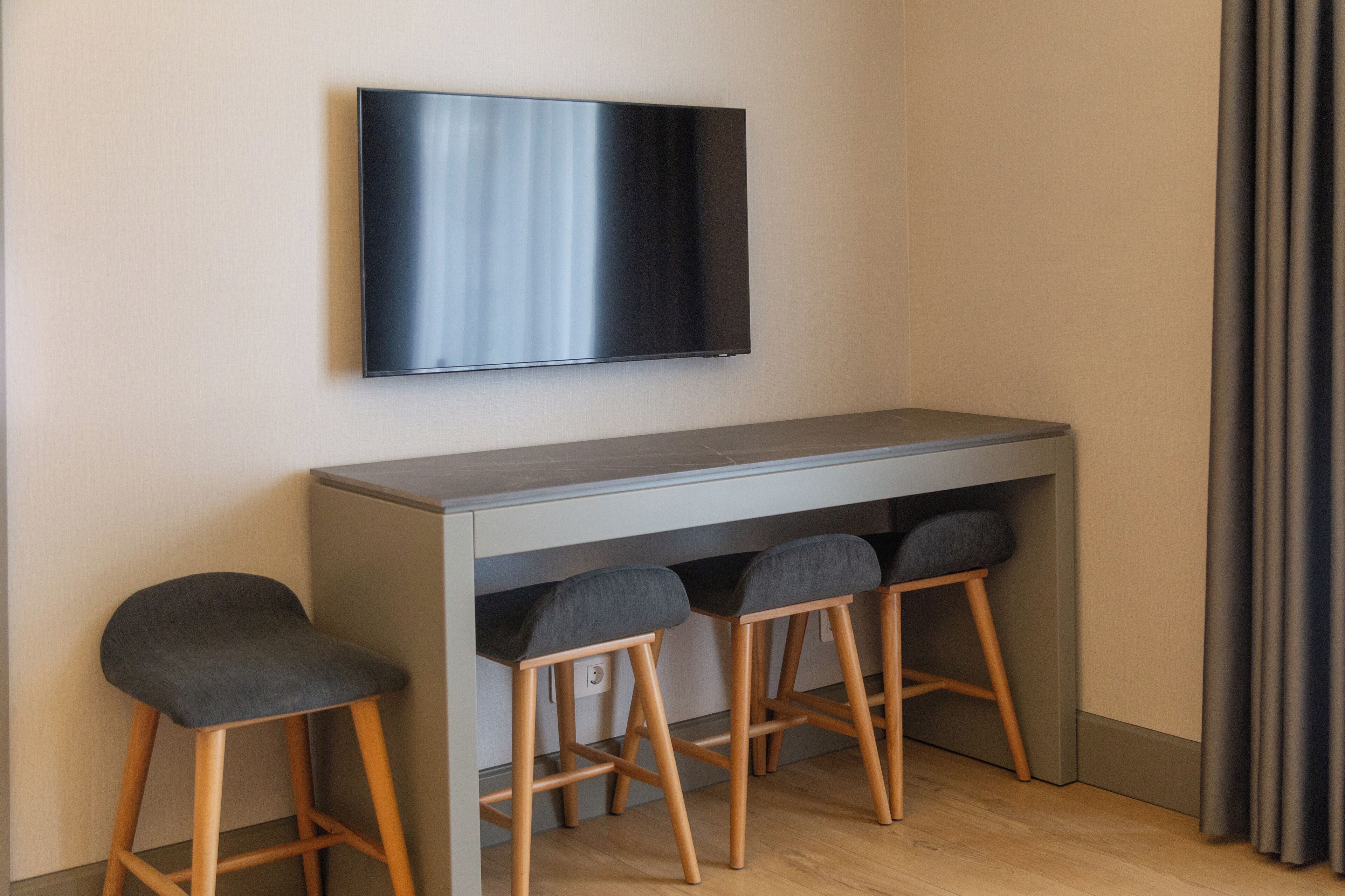 Quarto moderno com televisão de tela plana na parede, uma mesa cinza com dois banquinhos e uma janela com cortinas.