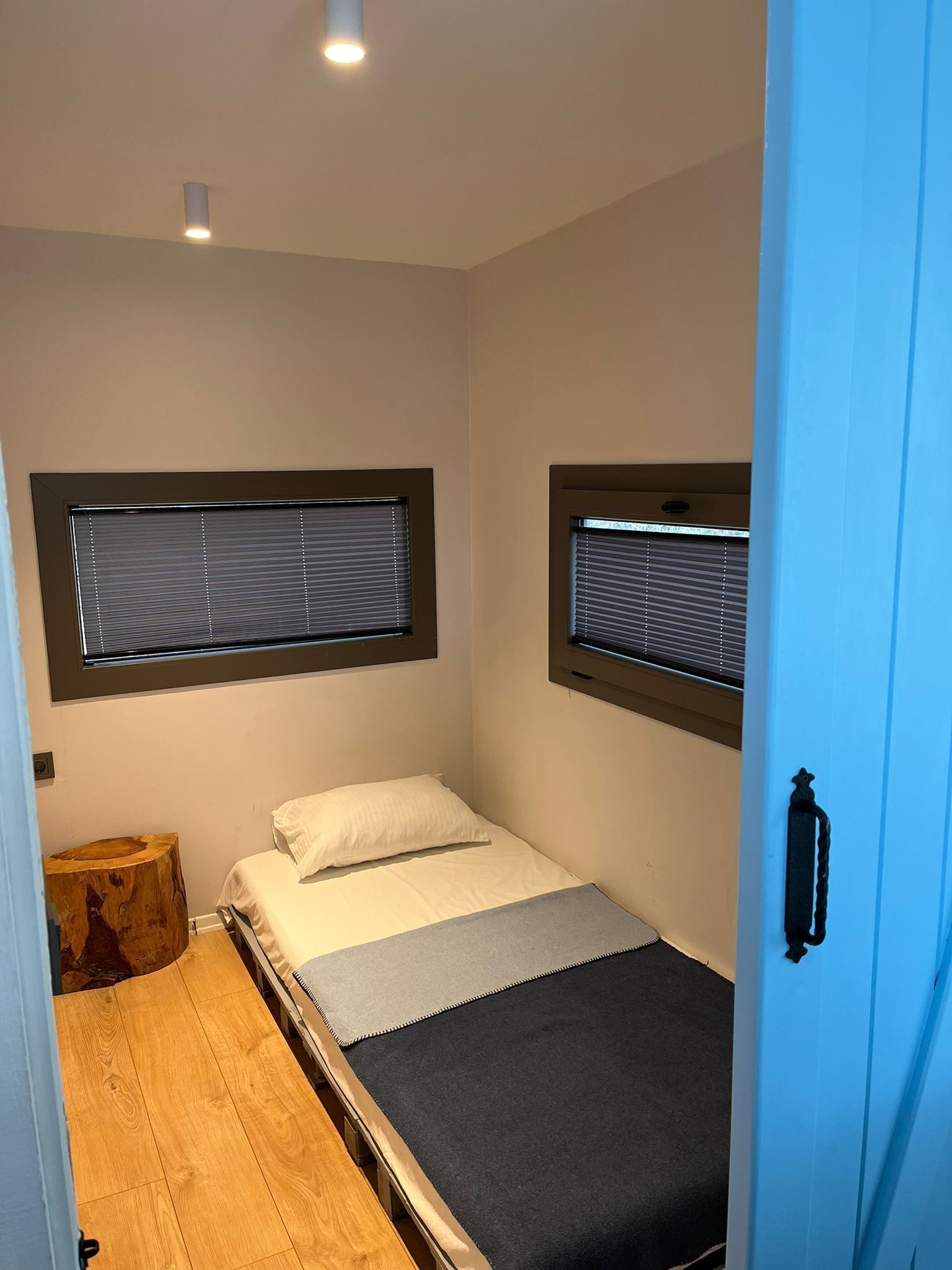 Quarto pequeno com cama de solteiro, piso de madeira, mesa de cabeceira e janela com persianas.