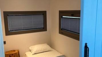 Quarto pequeno com cama de solteiro, piso de madeira, mesa de cabeceira e janela com persianas.