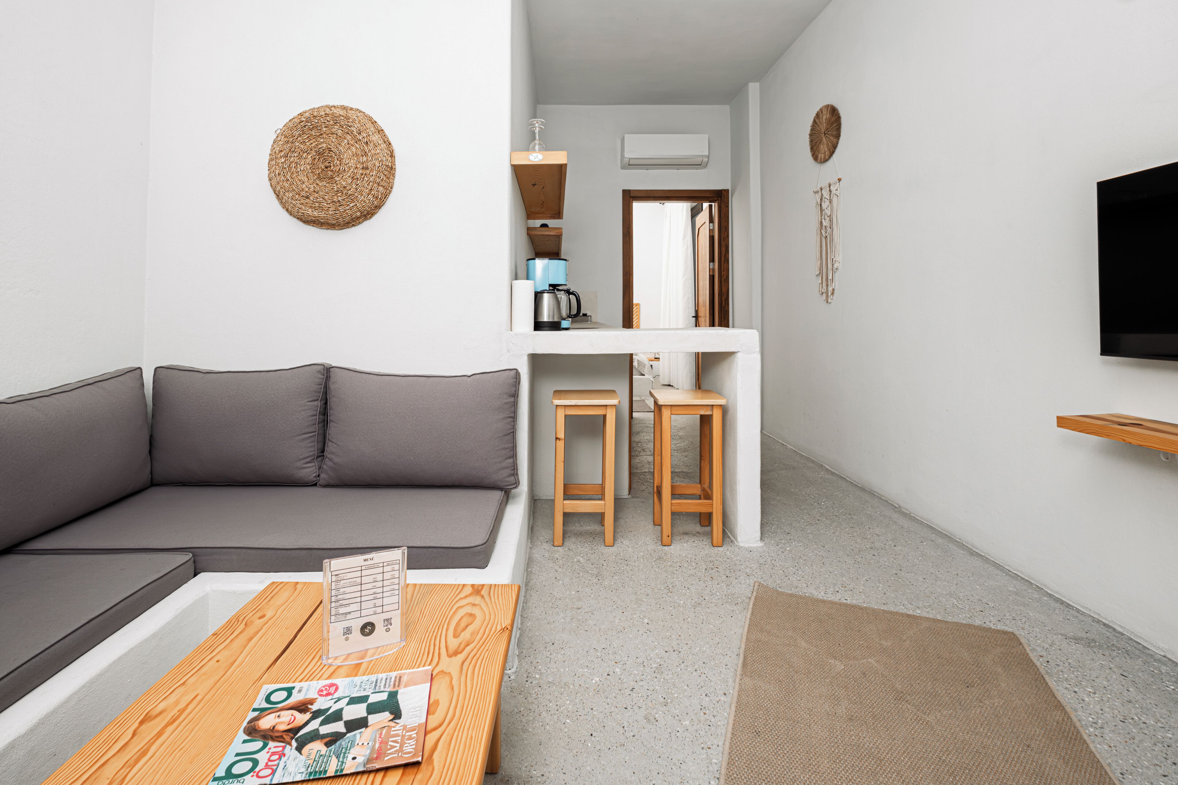 Una sala moderna con un sofá gris, una mesa de centro de madera y una revista. Hay una zona de cocina con taburetes y un televisor montado en la pared.