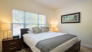 Une chambre à coucher comprenant un lit, deux tables de chevet, une commode, une fenêtre avec des stores et un tableau encadré accroché au mur.