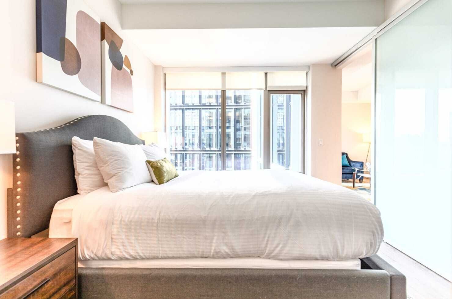A modern bedroom with a large bed, a nightstand, and a view of the city through large windows.
