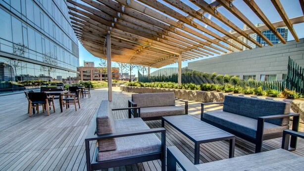 An outdoor seating area with wooden tables and chairs, a pergola structure, and a view of modern buildings.