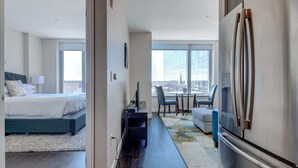 A hotel room with a bed, a desk, a chair, a refrigerator, and a view of the city.