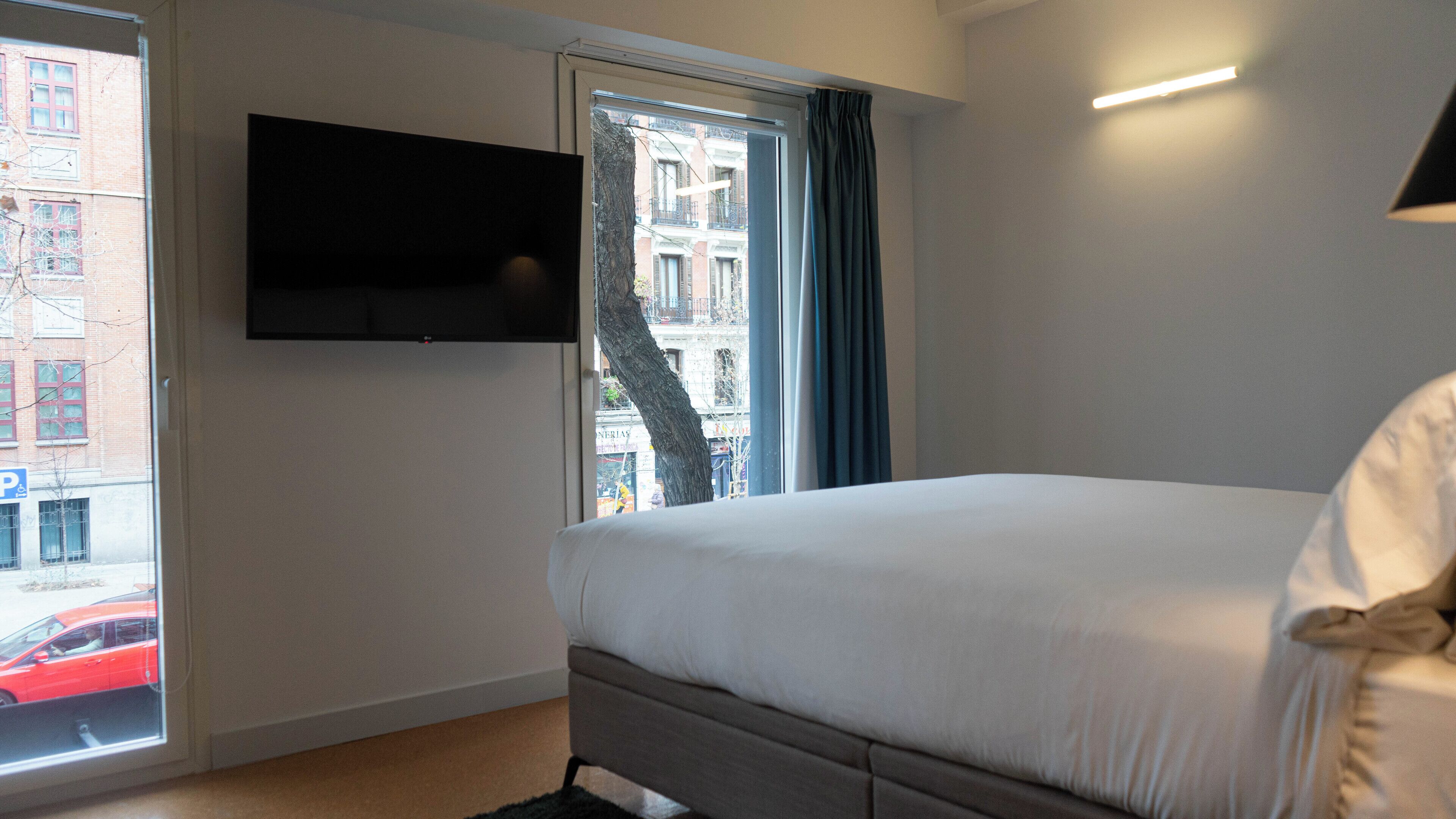Un dormitorio con una cama, un televisor montado en la pared y una ventana con cortinas.