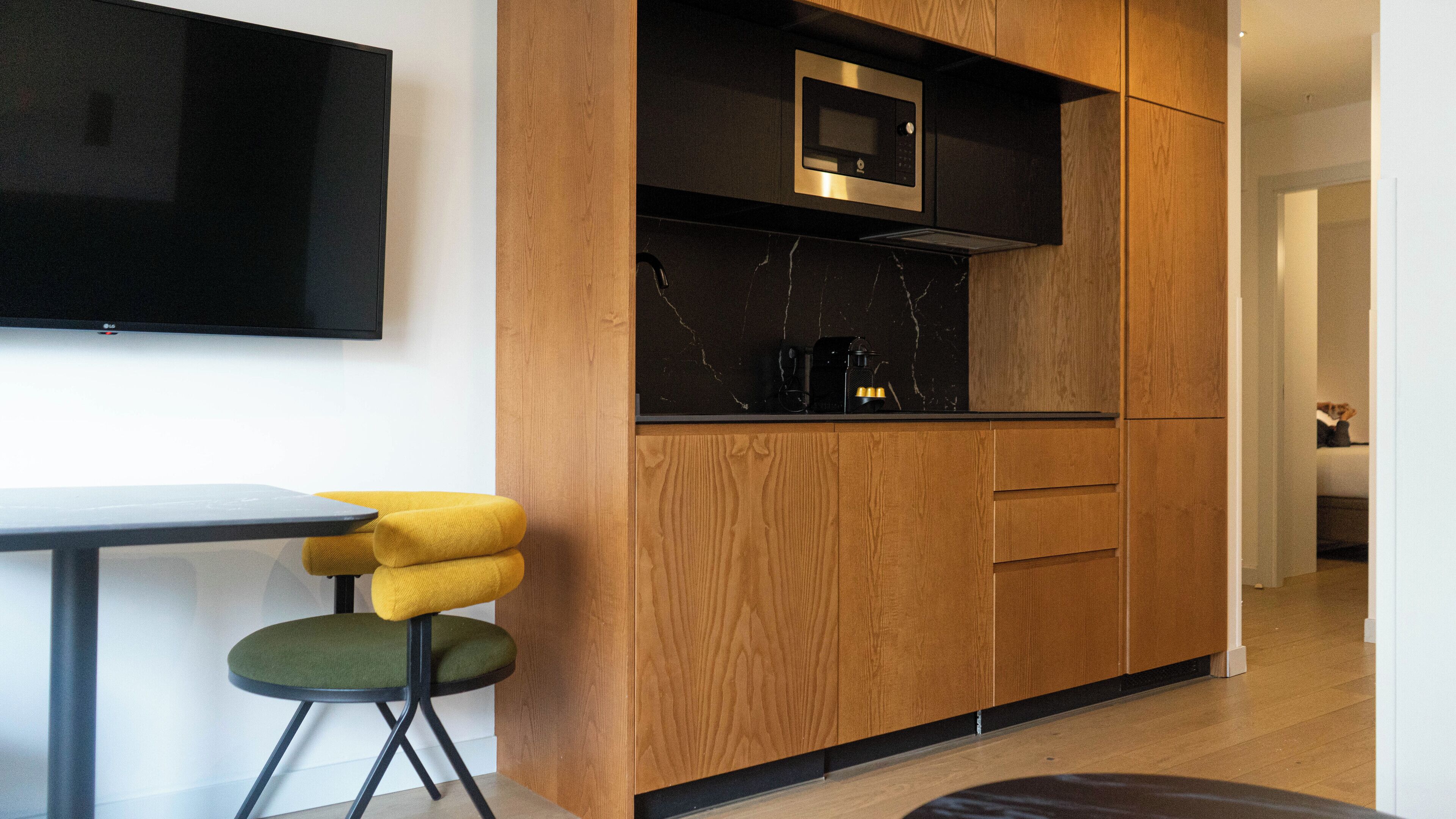A modern kitchen with wooden cabinets, a built-in microwave, and a small dining area with a yellow chair and a green stool.