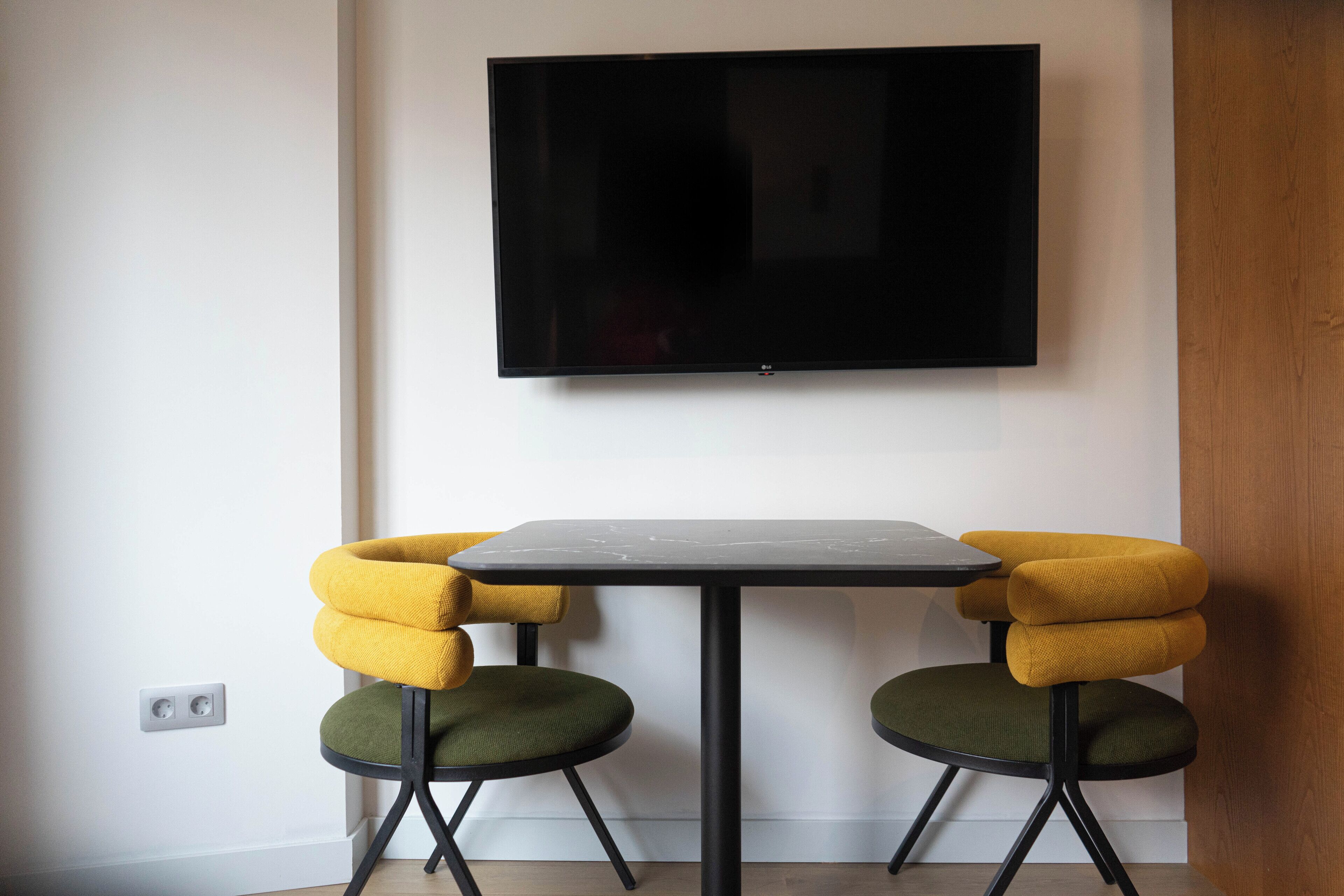 Una habitación moderna con una televisión de pantalla plana montada en la pared, una mesita pequeña y dos sillas con tapicería amarilla y verde.