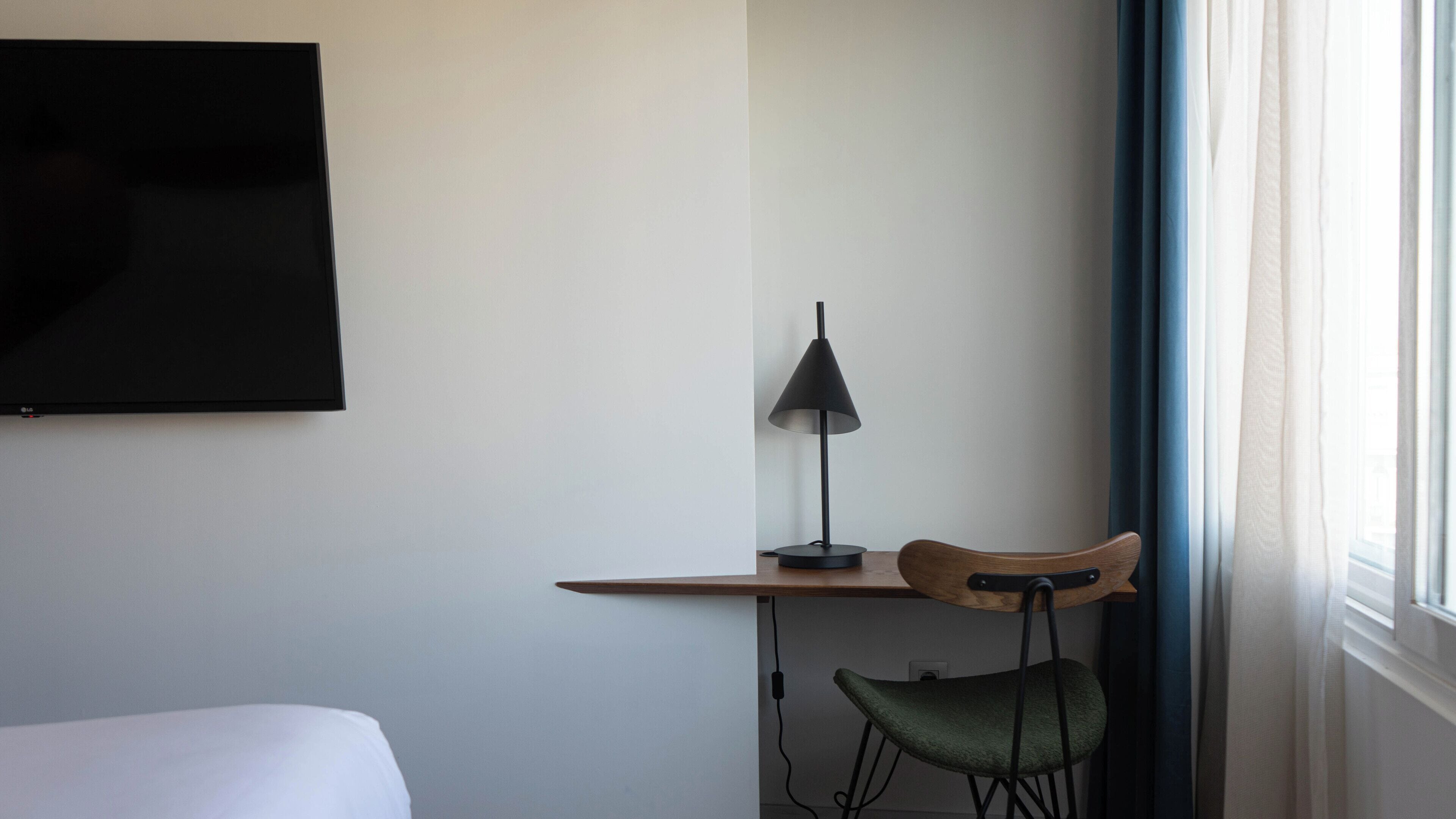 Ein Hotelzimmer mit einem Bett, einem Schreibtisch mit Lampe, einem Sessel und einem an der Wand befestigten Fernseher.