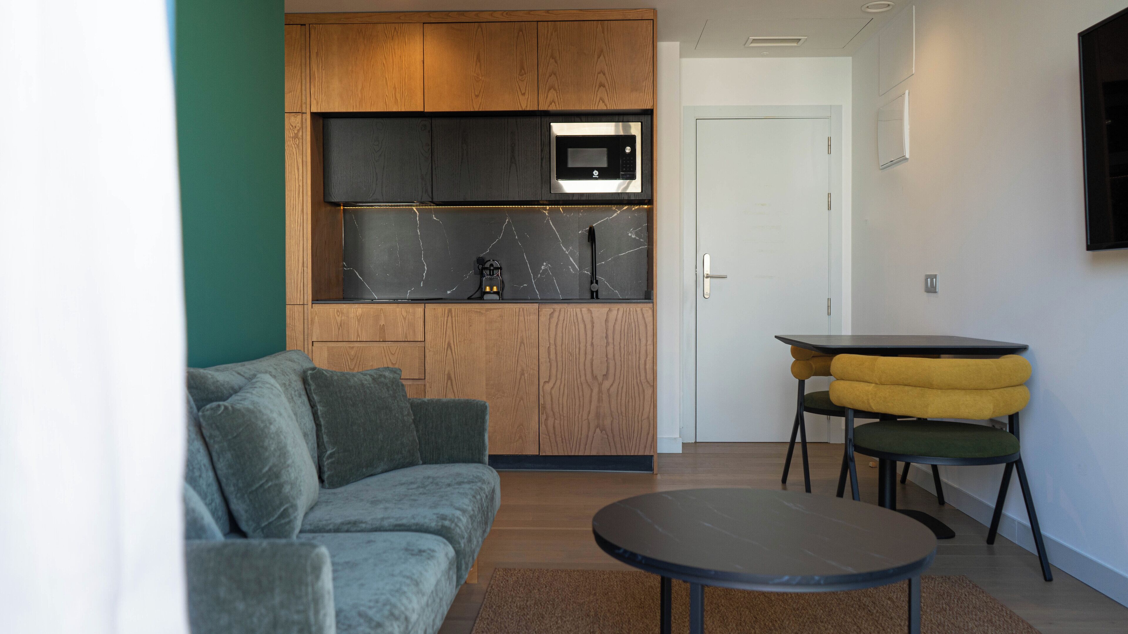 A modern living room with a grey sofa, a small round table, and a kitchen area with a microwave and wooden cabinets.