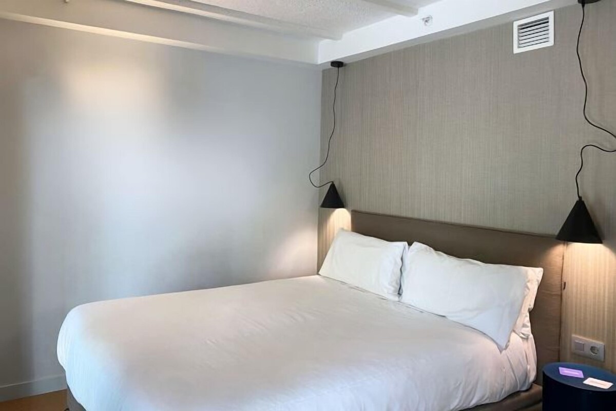 A neatly made bed with white linens in a room with a plain wall and a headboard with a light fixture.