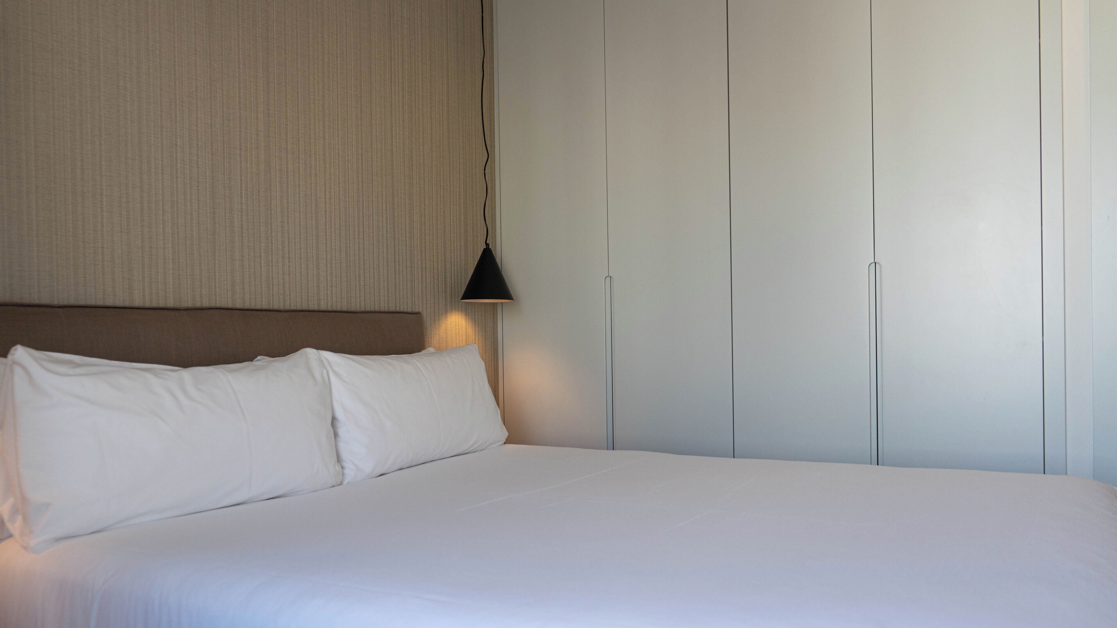 Hotellihuone, jossa on sänky, valkoiset vuodevaatteet, sängynpääty, seinään kiinnitetty lamppu ja vaatekaappi, jossa on pystysuorat vetimet.