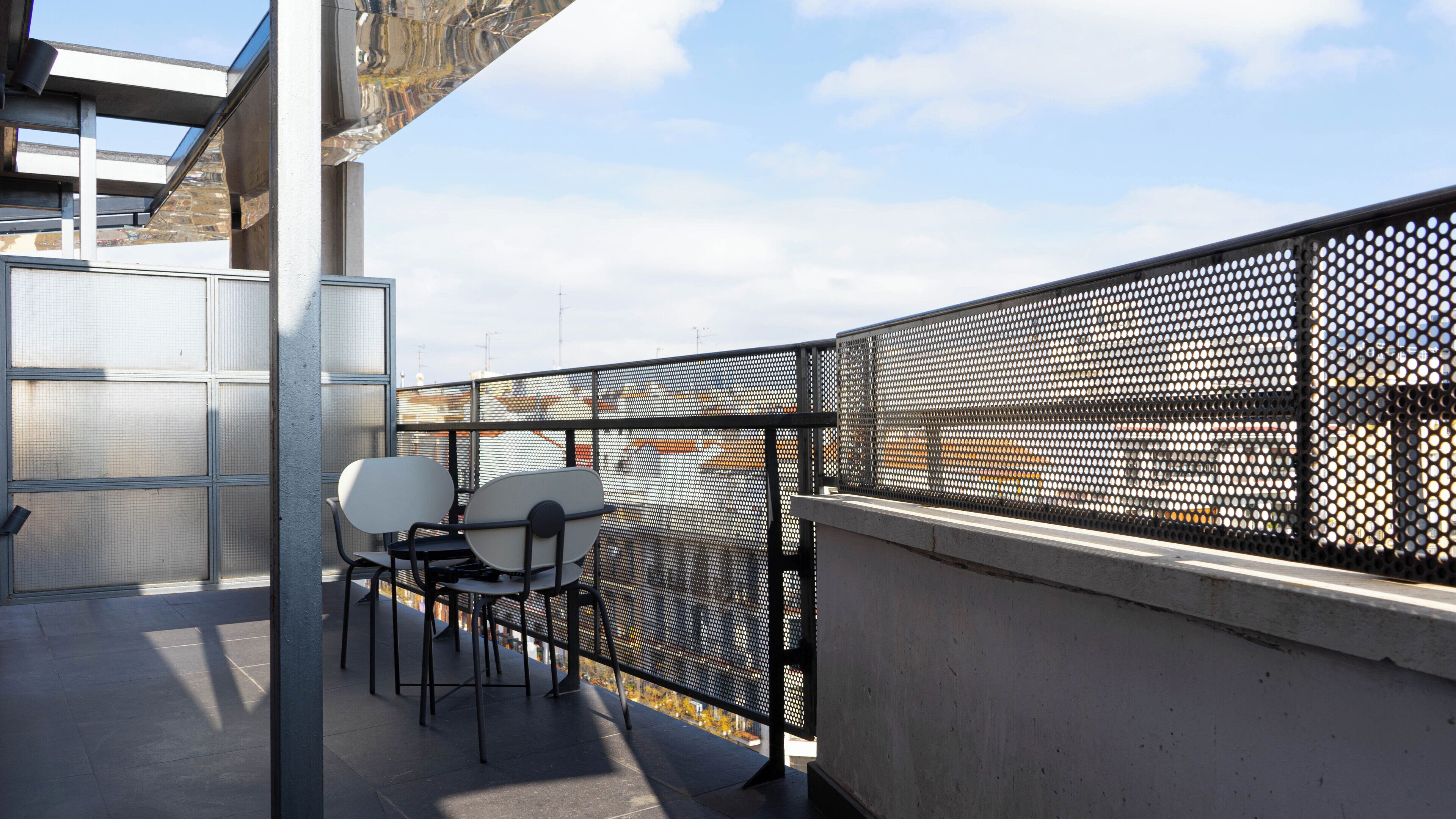 Un balcon avec une balustrade en métal, une table avec deux chaises et une vue sur des bâtiments en arrière-plan.