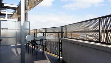 Un balcón con barandilla metálica, una mesa con dos sillas y vistas a edificios al fondo.